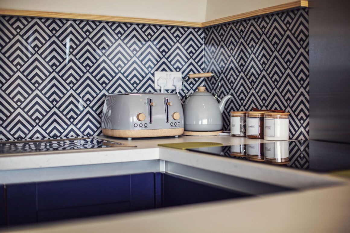 Modern kitchen corner with patterned backsplash, grey toaster, kettle, and tea, coffee, and sugar jars.